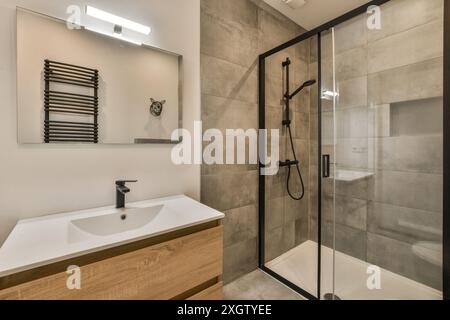 Elegantes, modernes Bad mit ebenerdiger Dusche, Kosmetikbereich aus Holz und minimalistischer Einrichtung mit neutralen grauen Fliesen. Stockfoto