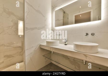 Das moderne Badezimmer verfügt über elegante weiße Marmorwände und Ablageflächen mit zwei stilvollen runden Waschbecken, modernen Armaturen und dezenter Beleuchtung. Stockfoto