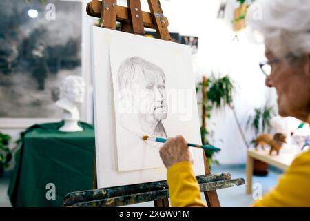 Eine ältere Frau, die sich auf das Malen eines männlichen Porträts konzentriert, arbeitet fleißig in ihrem pulsierenden Kunststudio, umgeben von anderen Kunstwerken Stockfoto