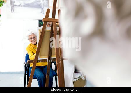 Eine ältere Frau mit einem fröhlichen Ausdruck malt auf einer Leinwand in einem hellen Atelier und zeigt künstlerische Leidenschaft und Kreativität in ihren älteren Jahren Stockfoto