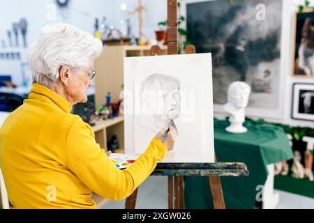 Eine ältere Frau mit weißen Haaren, die in einen gelben Pullover gekleidet ist, konzentriert sich auf das Malen eines Porträts auf Leinwand in einem gut beleuchteten Kunststudio, umgeben von verschiedenen Stockfoto