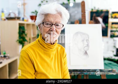 Eine ältere Frau mit einem hellen Lächeln, mit Brille und gelbem Rollkragen, steht neben ihrer Staffelei in einem gut beleuchteten Kunststudio und zeigt ein Complet Stockfoto