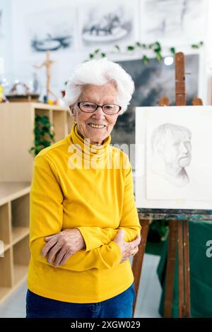Eine fröhliche ältere Frau mit weißem Haar und Brille, die in einem Kunststudio steht, posiert selbstbewusst vor einer Leinwand mit einer Bleistiftzeichnung. Tragen eines br Stockfoto