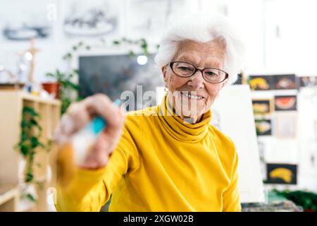 Ein älterer Künstler hält einen Pinsel und ein warmes Lächeln in einem hellen Kunststudio, umgeben von Gemälden Stockfoto