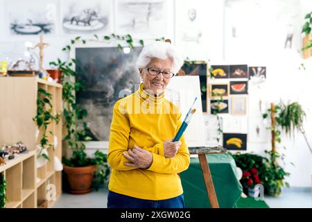 Eine fröhliche, ältere Künstlerin steht in ihrem Atelier, umgeben von Pflanzen und ihren Kunstwerken, mit Pinseln und einer hellgelben Turte Stockfoto