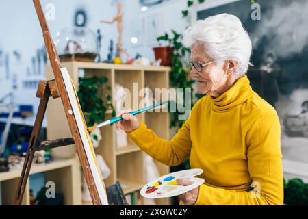 Eine ältere Frau in einem leuchtend gelben Pullover begibt sich mit der Malerei, hält eine Palette und einen Pinsel und konzentriert sich in einem kunstvollen Studio auf ihre Leinwand Stockfoto