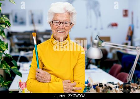 Eine ältere Frau mit weißen Haaren steht lächelnd in ihrem Atelier und hält einen Pinsel. Sie trägt einen hellgelben Rollkragen und eine Brille, umgeben Stockfoto