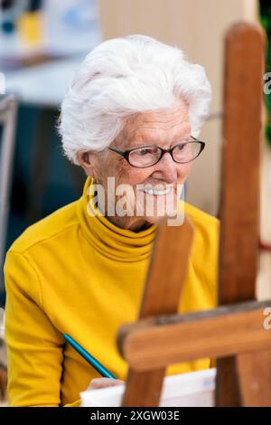 Eine fröhliche ältere Frau mit weißem Haar und Brille, trägt einen gelben Schildkrötenhals und malt an einer Staffelei in einem hellen Studio Stockfoto