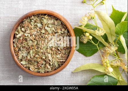 Linden blüht in einer Holzschale auf Leinenstoff. Getrocknete, frische Blüten, Früchte und Blätter der großblättrigen Linde oder Linde, Tilia platyphyllos. Stockfoto