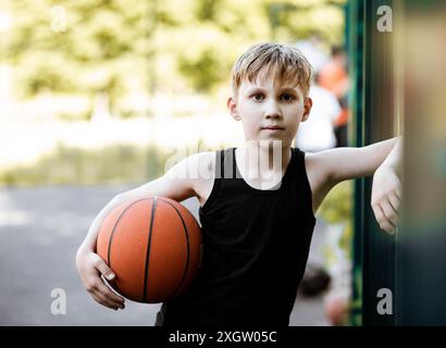 Ein kleiner Junge in einem schwarzen Tanktop steht selbstbewusst auf einem Basketballfeld im Freien und hält einen Basketball unter dem Arm. Der Hintergrund ist unscharf Stockfoto