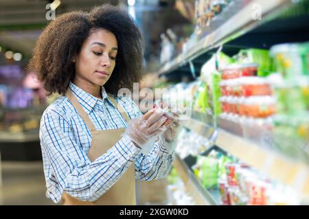 Supermarktmitarbeiter überprüft die Produktbeschriftung in den Regalen im Lebensmittelgeschäft. Weibliche Einzelhandelsarbeiterin mit Schürze und Handschuhen, die Lebensmittelverpackungen untersucht. Konzept der Qualitätskontrolle im Einzelhandel, Lebensmitteleinkauf Stockfoto