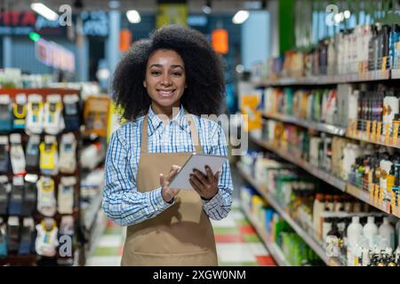 Weibliche Einzelhandelsangestellte, die im Ladengang ein Tablet hält, lächelt, Schürze und kariertes Hemd trägt. Mitarbeiter demonstrieren einen modernen Ansatz für Bestandsverwaltung und Kundenservice Stockfoto