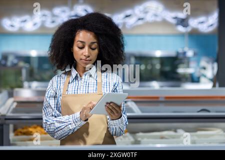 Arbeiter in der Schürze mit Tablette in der Fischabteilung im Supermarkt. Das Bild zeigt eine junge Frau, die mit moderner Technologie Inventar überprüft. Konzept für Einzelhandel, Meeresfrüchte, Fisch und Lebensmittelgeschäft. Stockfoto