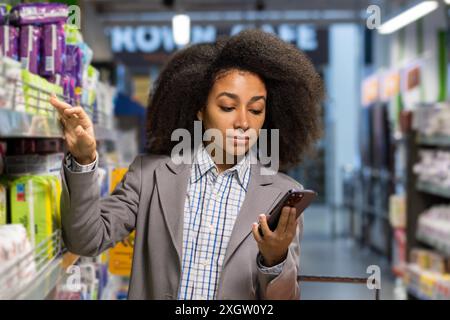 Junge Frau, die im Supermarkt einkaufen, während sie ihr Handy überprüft. Sie trägt einen Business-Anzug und sammelt Gegenstände aus dem Regal. Multitasking-Konzept und moderner Lebensstil. Stockfoto