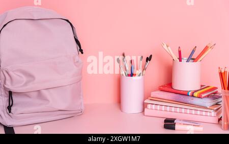 Lila Rucksack auf einem Tisch mit Schulutensilien vor rosa Hintergrund Stockfoto