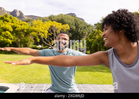 Ein birassisches Paar genießt einen lustigen Moment im Freien beim Yoga. Sie lachen, während sie sich in Yoga-Posen in einem sonnigen Garten ausstrecken. Stockfoto