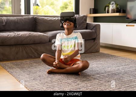 Der afroamerikanische Junge genießt die virtuelle Realität zu Hause. Er ist in eine digitale Welt eingetaucht und sitzt im Kreuz auf dem Wohnzimmerboden. Stockfoto