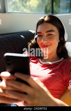 Das kaukasische Teenager-Mädchen genießt zu Hause Musik auf ihren hübschen Kopfhörern. Sie entspannt sich auf einer Couch, verlobt mit ihrem Smartphone in einem sonnendurchfluteten Raum. Stockfoto