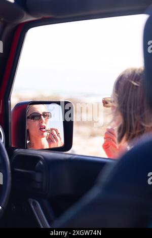 Die junge kaukasische Frau trägt Lippenstift im Autospiegel auf einer Autofahrt, mit Kopierraum. Die Momente sind eine Mischung aus Schönheitsroutine und Reisen. Stockfoto