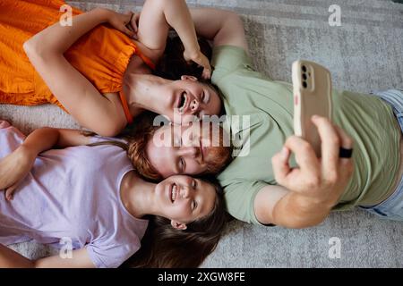 Drei Freunde liegen auf dem Rücken auf dem Boden, ihre Köpfe bilden einen Kreis. Der Mann in der Mitte hält ein Smartphone und macht ein Selfie Stockfoto