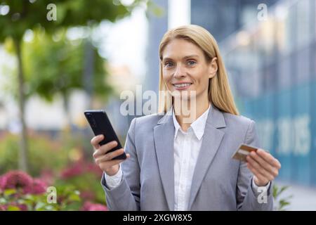 Selbstbewusste Geschäftsfrau im grauen Anzug, die ihr Smartphone für Online-Zahlungen nutzt, während sie ihre Kreditkarte hält. Im Freien in urbaner Umgebung mit modernen Gebäuden im Hintergrund. Stockfoto