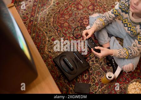 Abgeschnittener Blick von oben auf den kaukasischen Teenager in gemustertem Pullover und Jeans mit einem Retro-Video-Game-Controller, während er auf einem Vintage-Teppichboden sitzt, neben einer Drink- und Popcorn-Schüssel Stockfoto