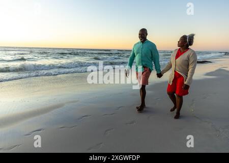 Ein älteres afroamerikanisches Paar genießt einen Strandspaziergang bei Sonnenuntergang. Ihr entspannter Spaziergang entlang der Küste fängt einen Moment des ruhigen Ruhestands ein. Stockfoto