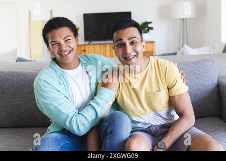 Junger birassischer Mann und Mann aus dem Nahen Osten sitzen auf einer Couch zu Hause während eines Videoanrufs, mit Kopierraum. Sie teilen einen freudigen Moment, reflektieren ein Co Stockfoto