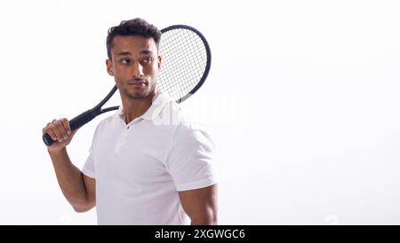 Junger birassischer Mann posiert mit einem Tennisschläger auf weißem Hintergrund, mit Kopierraum. Er steht zuversichtlich in einem Studio-Setting und schlägt einen aktiven vor Stockfoto