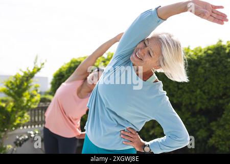 Zwei glückliche ältere Freundinnen üben Yoga im Stehen und Arm Strecken im sonnigen Garten. Yoga, Wohlbefinden, Bewegung, Freundschaft und Gesundheit Stockfoto