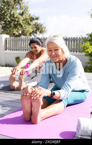 Zwei verschiedene ältere Freundinnen, die Yoga machen und Beine dehnen, sitzen im sonnigen Garten, Kopierraum. Yoga, Wohlbefinden, Bewegung, Freundschaft und gesunde Geselligkeit Stockfoto