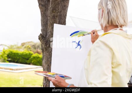 Ältere kaukasische Frau malte an der Staffelei im sonnigen Garten, Kopierraum. Sommer, Kunst, Kreativität, Hobbys, Ruhestand und aktive seniorenleben Stockfoto