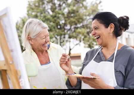 Zwei glückliche ältere Freundinnen, die sich an der Staffelei im Garten unterhalten und malen. Freundschaft, Kunst, Kreativität, Hobbys, Ruhestand und aktive Seniorinnen Stockfoto