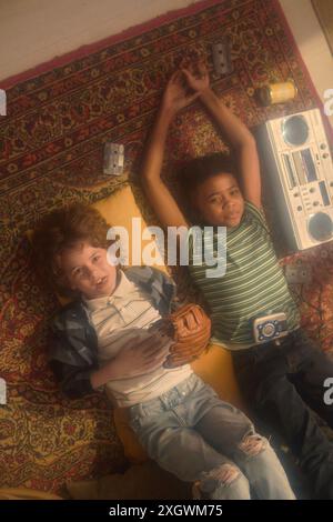 Top-Down-Aufnahme des afroamerikanischen Jungen und des kaukasischen Jungen, der auf gemustertem Teppich mit Boombox, Kassetten und Baseballhandschuh liegt und entspannte Momente im Retro-Ambiente genießt Stockfoto