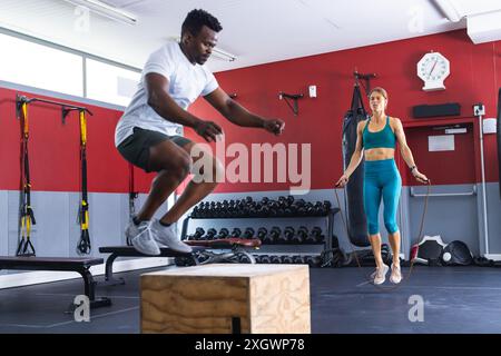 Fitter afroamerikanischer Mann springt im Fitnessstudio auf eine Box, fitte junge kaukasische Frauen trainieren mit einem Seil. Sie konzentrieren sich auf ihr intensives Workout Stockfoto