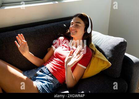 Das kaukasische Teenager-Mädchen entspannt sich zu Hause, hört Musik auf ihren hübschen Kopfhörern und sonnt sich im Sonnenlicht. Stockfoto