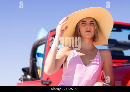 Die junge kaukasische Frau schirmt ihre Augen vor der Sonne auf einem Roadtrip ab. Sie steht draußen in der Nähe eines roten Autos, trägt einen stylischen Hut und ein Sommerkleid. Stockfoto