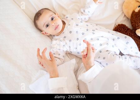 Lächelndes Baby im Pyjama mit Sternenmuster, das mit einem Teddybären auf einem weißen Bett liegt und liebevoll von einem Elternteil betreut wird. Ideal für Stockfotos auf Eltern Stockfoto