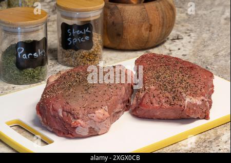 Zwei rohe Steaks mit Gewürzen bereit zum Barbeque auf weißem Schneidebrett Nahaufnahme. Holzschale und Gewürze im Hintergrund. Stockfoto