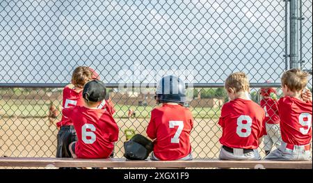 Eine Reihe junger T-Ball-Jungs, die auf der Bank hinter dem Heimteller sitzen und darauf warten, zu schlagen. Stockfoto