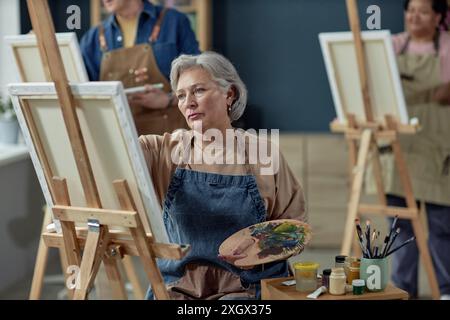 Porträt einer grauhaarigen Seniorin, die auf Staffelei malte und Künstlerpalette im Kopierraum des Kunststudios hielt Stockfoto