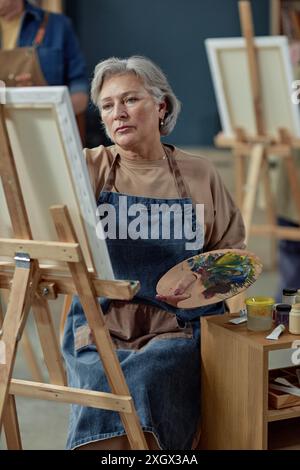 Vertikales Porträt einer eleganten kaukasischen Seniorin, die ein Bild auf Staffelei im Kunststudio malte und eine Palette mit Ölfarbe hielt Stockfoto