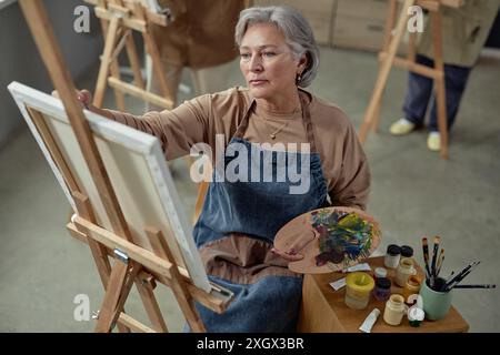 Hochwinkel-Porträt einer kreativen Reifen Frau, die im Kunststudio ein Bild auf Staffelei malt und eine Palette hält, die Inspiration sucht Stockfoto