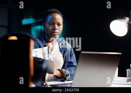 Schwarze Frau mit Papierkram zum Vergleich mit Informationen auf dem PC. Engagierte afroamerikanische Dame, die online recherchiert und studiert, mit ihrem digitalen Laptop in ihrer Wohnung. Stockfoto
