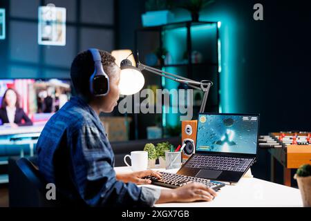Afroamerikanerin spielt ein kompetitives Videospiel in ihrem gemütlichen Wohnzimmer mit einem kabellosen Computer und Kopfhörern. Junge Dame mit Tastatur, Maus und Laptop für Online-Spiele. Stockfoto