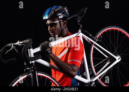 Afroamerikaner hält ein Rennrad auf schwarzem Hintergrund, mit Kopierraum. Er trägt Fahrradausrüstung und ist bereit für ein Rennen oder Training. Stockfoto