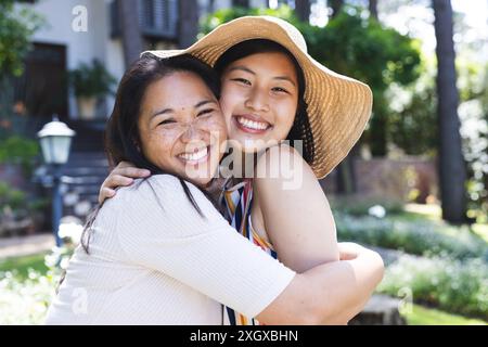 Zwei asiatische Frauen, eine Mutter und eine Tochter, teilen sich eine warme Umarmung im Freien, mit Kopierraum. Ihr fröhlicher Ausdruck und ihre enge Umarmung vermitteln eine starke Bindung Stockfoto
