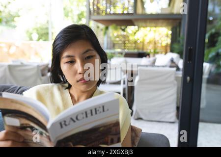 Fokussierte asiatische Frau auf Couch, die zu Hause Buch liest. Leben, Literatur und Lebensstil unverändert. Stockfoto