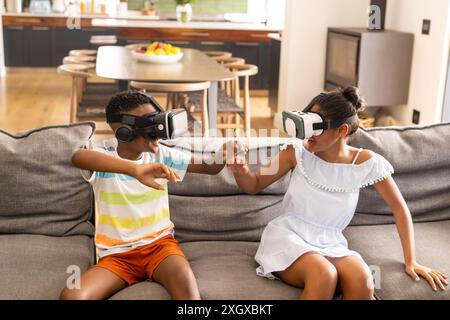 Birassische Geschwister genießen VR-Spiele zu Hause. Sie haben Spaß mit Virtual-Reality-Technologie in einem gemütlichen Wohnzimmer. Stockfoto