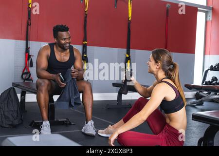 Fitter afroamerikanischer Mann plaudert mit einer jungen, fitten Kaukasierin im Fitnessstudio. Sie teilen einen leichten Moment während einer Trainingspause und fördern Fitness und CA Stockfoto